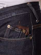 Close-up of a person wearing Repi Dai V Dark Jeans by Blue de Gênes in dark blue denim. Focus on the pocket area, featuring gold stitching and a metal button. The regular slim fit shows comfort stretch fabric with slight fading and frayed threads near the pocket top.