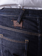 Close-up of Repi Dai V Dark Jeans by Blue de Gênes in dark blue comfort stretch denim, showcasing gold stitching and a cow-embossed brown leather patch. The regular slim-fit jeans are worn with a hint of a white shirt visible at the waistband.
