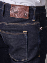Close-up of a person wearing Blue de Gênes Repi Dai V Dark Jeans in dark blue comfort stretch denim, featuring a brown leather patch on the waistband labeled Blue de Gênes. Gold stitching and a small decorative patch with a cross design on the pocket highlight Repi Jeans attention to detail.