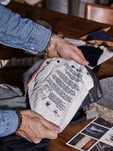 A person stylishly holds the interior of a denim jacket showcasing printed text in multiple languages. They sport a blue denim shirt and bracelets, echoing the effortless style of Blue de Gênes Repi Dai V Dark Jeans in dark blue denim. A wooden table with papers and fabric is visible in the background.