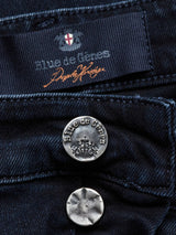 Close-up of the Vinci 3325 Black Jeans from Blue de Gênes reveals decorative writing on the label. Below, two metal buttons feature Blue de Gênes engraving and a coat of arms. These expertly crafted Italian jeans in deep navy Comfort Stretch Denim promise both style and ease.