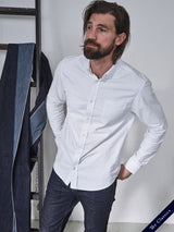 A man with a beard, in a Sandro Oxford Shirt - White and dark jeans, stands by a white wall. He has long, dark hair and is near a ladder draped with blue and denim fabric. A label reads Blue de Gênes: The Classics.