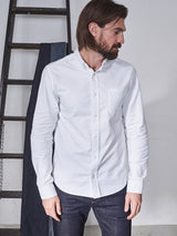 A man with brown hair and a beard, wearing the BDGsandro Oxford Shirt in white by Blue de Gênes and dark jeans, stands near a wooden ladder against a light wall, looking to his left.