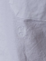 Close-up of the Blue de Gênes BDGsandro Oxford Shirt in white, featuring a visible shield-shaped patch repair and a diagonal seam across the upper fabric.