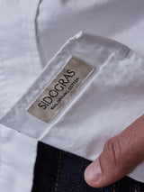 A close-up of a white clothing label reading Blue de Gênes - LINEN ORIGINAL COTTON sewn onto the BDGsandro Oxford Shirt in White, with a hand holding the edge and dark blue denim below—showcasing perfect detailing.