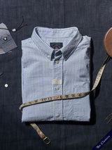 A folded Sandro Oxford Shirt - Blue Stripe by Blue de Gênes is placed on dark fabric, surrounded by a measuring tape, a tag, a metal button, and a small watch. A corner tag reads The Classics.