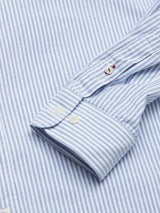 Close-up of the Sandro Oxford Shirt by Blue de Gênes, featuring blue and white stripes, buttoned cuffs with red and white buttonhole stitching detail. This organic cotton fabric offers a subtle texture for an elegant design.
