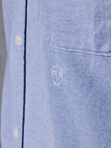 A close-up of the Sandro Oxford Shirt - Blue by Blue de Gênes shows a light blue shirt made from organic cotton, featuring an embroidered shield emblem on the chest pocket. The subtly textured fabric reveals two white buttons on the placket.