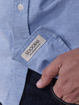 Close-up of a person wearing the Sandro Oxford Shirt - Blue, featuring a pocket. A beige tag reads SIDO GRAS 100% ORIGINAL COTTON. This Blue de Gênes button-down complements the persons hand tucked into the pocket of dark blue jeans.