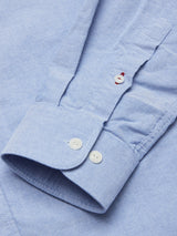 Close-up of the sleeve of a Sandro Oxford Shirt - Blue by Blue de Gênes, made from textured organic cotton. It features two white cuff buttons and a red placket button, with slightly wrinkled yet stylish fabric.