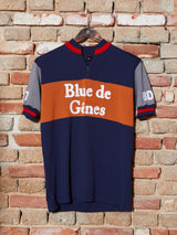 The Tributo Cycling Jersey - Navy by Blue de Gênes, crafted from Merino Wool, features blue, orange, and gray hues. With the brands name on the front and numbers 87 and BD on the sleeves, it hangs vintage-style on a wooden hanger against a brick wall—a classic cycling piece.