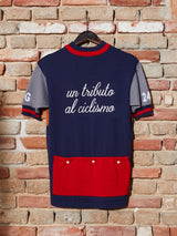 A vintage-style Tributo Cycling Jersey by Blue de Gênes hangs on a brick wall, crafted from premium Merino Wool. Featuring short sleeves, a navy body with gray sleeves, red collar/hem, and un tributo al ciclismo in white script on the back above two red pockets.