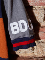 Close-up of a grey fabric with BDG embroidered in white, similar to Blue de Gênes Tributo Cycling Jersey. The edge has red and blue stripes, like cycling attire. Background is a textured brick wall.