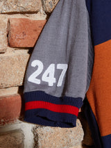 A close-up of a sleeve against a brick wall, resembling the Tributo Cycling Jersey by Blue de Gênes. The gray sleeve with white 247 embroidery features Merino Wool elements, transitioning from a blue cuff with red stripes into a navy and orange body, echoing classic cycling styles.