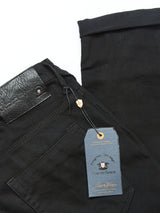 Close-up of Repi Stay Black Jeans by Blue de Gênes, featuring a leather label and gray tag with brand details and a crossed flag emblem. Made from comfort stretch denim, the black slim-fit jeans are neatly folded against a white background.