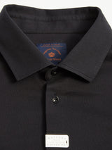 A close-up of the Luccio Dry Shirt - Jet Black from Blue de Gênes showcases a neatly folded polo crafted from cotton pique jersey with garment-dyed fabric, a collar, and button details. A small tag reads: Buttons: Ecological Corozo Nut, highlighting its quality craftsmanship.