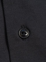 Close-up of the Luccio Dry Shirt - Jet Black by Blue de Gênes, showcasing its dark, textured cotton pique jersey. A small, shiny metallic button inscribed with rue de Classe is sewn on, reflecting the finely woven fabric and hinting at garment-dyed craftsmanship.