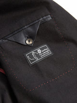 Close-up of the Leopardi Carbonio Blazer by Blue de Gênes, showcasing black fabric with red stitching and buffalo horn buttons. A buttoned black flap above the pocket features a label saying “Lanificio F.lli Cerruti, Woven in Italy.”.