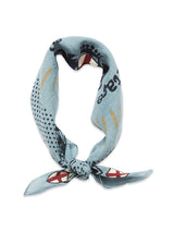 The Blue de Gênes Bandana - Celeste is a stylish cotton neck scarf in blue, featuring polka dots and abstract patterns in black and light brown, along with red and white shield-like designs. It is folded into a loop with loosely knotted ends.