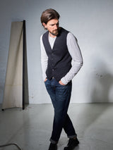 A bearded man with medium-length hair stands contemplatively, his Italian brushed cotton white long-sleeve shirt visible under a Blue de Gênes Canna Carbonio Gillet in black with Buffalo horn buttons. He pairs this with dark jeans and black shoes, set against the minimalistic backdrop of a white-walled room.