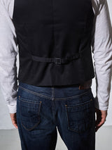 The image shows a person from behind wearing a white long-sleeve shirt, blue jeans, and the Blue de Gênes Canna Carbonio Gillet in black. The vest features buffalo horn buttons, crafted from Italian brushed cotton with an adjustable back strap and a visible pocket clip.