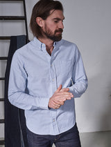A bearded man in a Blue de Gênes Sandro Oxford Shirt with blue stripes and dark jeans stands near a ladder. He gazes to the side, hands clasped, in a minimalistic setting with a plain background, reflecting simple elegance and subtle sophistication.