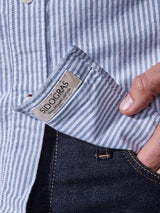 Close-up of the Sandro Oxford Shirt - Blue Stripe by Blue de Gênes, made from organic cotton, being slightly tugged from dark jeans. A hand is partially visible holding the shirt.