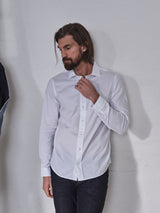 A long-haired, bearded man stands thoughtfully against a white wall, wearing the Miguel Brilliante Shirt - White by Blue de Gênes. Made of Italian cotton with Corozo nut buttons, his dark jeans complete the look as he holds the shirt collar and gazes to the side.