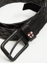 The Intero Belt - Black by Blue de Gênes is a black braided leather belt with a matte black buckle, hand-sewn stitches, and features a small red cross emblem on a white background stitched near the buckle.