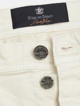 Close-up of ecru Paulo Pavia 1 Chino by Blue de Gênes featuring two metallic buttons engraved with the brand name. Above them, a dark label displays Blue de Gênes and a logo. Visible stitching highlights craftsmanship, while the cotton stretch fabric ensures both comfort and style.