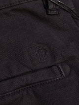 Close-up of the Teo Chino - Jet Black by Blue de Gênes, featuring dark garment dyed honeycomb fabric and a stitched shield-and-cross emblem above a neatly sewn pocket—a refined accent on classic chinos.