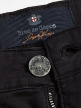 Close-up of Teo Chino - Jet Black pants by Blue de Gênes, featuring a metal button with the brand crest, visible zipper, and inner label displaying the brand name with an orange signature.