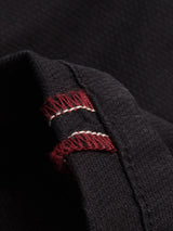 Close-up of Blue de Gênes Teo Chino - Jet Black, showing garment dyed fabric with visible texture and a folded edge. Red and white stitching detail stands out along the seam.