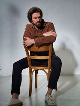 A man with medium-length hair and a beard sits backward on a wooden chair, wearing Blue de Gênes Repi 3325 Black Jeans in black denim, a brown jacket, and white sneakers against a plain light background.