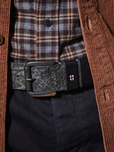 Close-up of a person wearing a brown corduroy jacket, plaid shirt, and dark pants. The focus is on the textured Piceno Leather Belt by Blue de Gênes in black cow leather with a metal buckle featuring a white cross on a red shield for a distinctly vintage look.