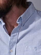 A close-up shows a person in the Blue de Gênes Sandro Oxford Shirt - Blue Stripe, with a vertically striped pattern. The top button is undone, and theres a pocket on the left side. Made from organic cotton, only part of the individuals lower face and neck is visible.