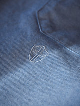 Close-up of a blue textured fabric, like the Sandro Oxford Shirt by Blue de Gênes, showing a small white shield emblem with a symbol. Featuring a subtle grid pattern and an angled seam in the upper right corner, made from soft organic cotton.
