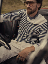 In a vintage car, a man sports Blue de Gênes sunglasses, a striped long-sleeve shirt, scarf, and Paulo Pavia 1 Chino - Ecru by Blue de Gênes. His bearded, relaxed demeanor is evident as he rests one hand on the wheel with grassy fields stretching out behind him.