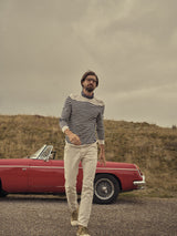 A man with glasses and a beard confidently walks in front of an open-top red vintage car, wearing a striped long-sleeve shirt, Paulo Pavia 1 Chino - Ecru by Blue de Gênes, and beige shoes. The backdrop features a grassy hill under a cloudy sky.