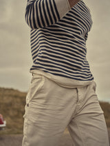 A person is outdoors, wearing a navy and white striped sweater paired with Blue de Gênes Paulo Pavia 1 Chino in Ecru. The focus is on the cotton stretch fabric of the chinos, set against a blurred grassy background with an overcast sky and a red car.