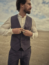 A man with a beard and long hair stands in a barren landscape under a cloudy sky, wearing a white shirt, trousers, and the Blue de Gênes Canna Pallone Gillet - Navy Stripe. He adjusts his cotton linen vest with hands adorned with rings.