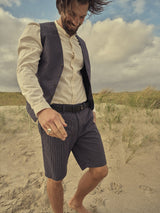 A man with long hair and a beard smiles, walking barefoot on a sandy beach. He wears a light cotton linen shirt, the Canna Pallone Gillet - Navy Stripe by Blue de Gênes, and matching pinstripe shorts. The sky is cloudy, with grasses growing in the sand.