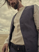A man with tousled hair stands outdoors under a cloudy sky, wearing a crisp white shirt and a Canna Pallone Gillet - Navy Stripe by Blue de Gênes. His gaze tilts downward as one hand rests in his pocket, enhancing the moody, vintage feel of the cotton linen.
