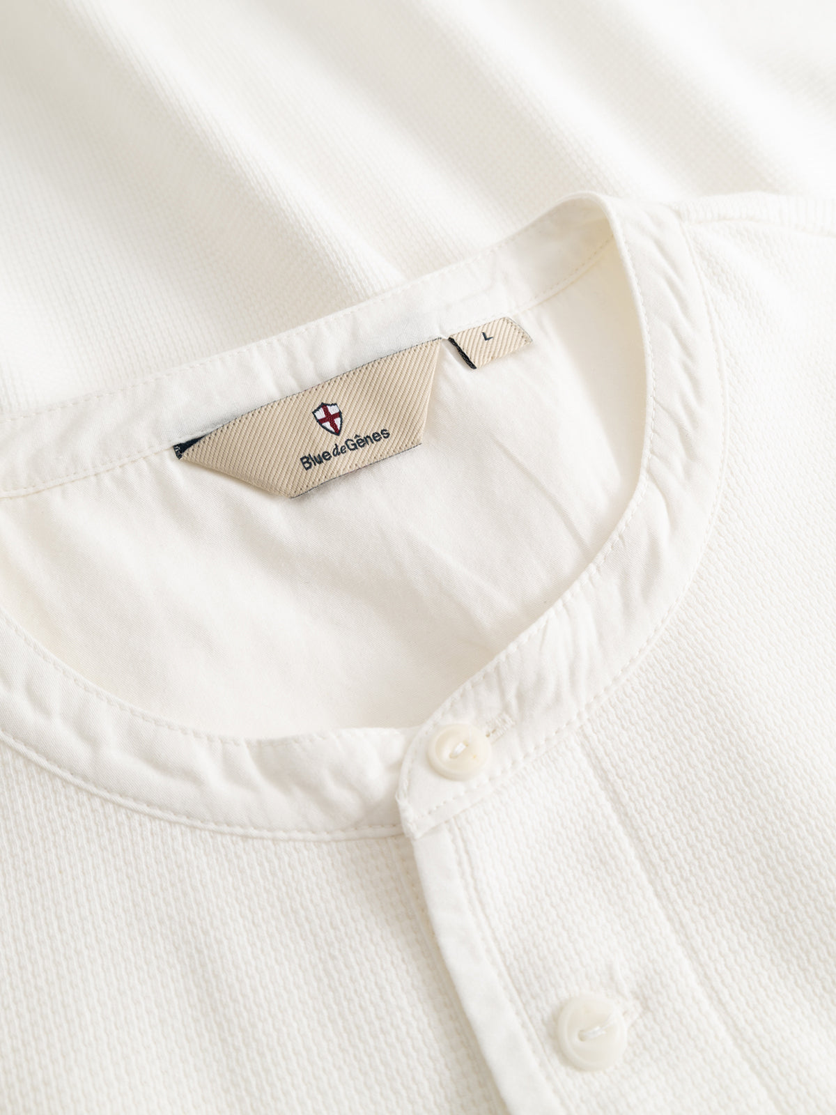 Close-up of a BGgreg L/S Grandad shirt by Blue de Gênes: white cotton poplin with a round collar, corozo nut buttons, and an inner fabric tag showing the Blue de Gênes logo in red and blue.