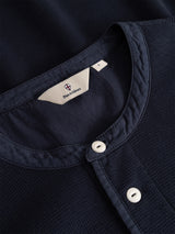 Close-up of the BGgreg L/S Grandad shirt by Blue de Gênes in dark navy, featuring a regular fit, round collar, white corozo nut buttons, and textured cotton fabric. The inner label is stitched near the neckline.