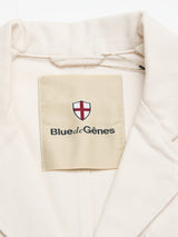 A close-up of the BGbaio Twill Blazer - Chalk highlights its refined beige collar and the Blue de Gênes label beneath a red and white shield logo, showcasing the elegant details of this classic cotton blazer.