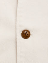 Close-up of a brown four-hole button, featuring small engraved text, sewn onto off-white ribbed fabric near a seam—showcasing the refined detailing of the Blue de Gênes BGbaio Twill Blazer in Chalk.