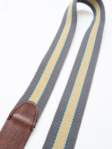 A close-up of the Blue de Gênes BGcollege Belt - Green Stripe shows green-striped webbing and a brown leather end piece in the lower left corner.
