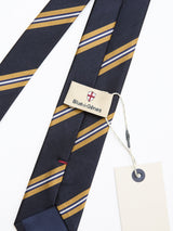 The BGponsy Tie - Dark Navy by Blue de Gênes is a navy blue silk tie with yellow and white diagonal stripes in a style reminiscent of classic khaki-striped ties, featuring a Blue de Gênes tag and a blank white price tag attached with string.