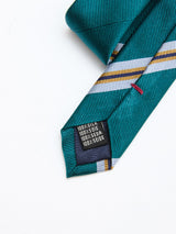 A closeup of the end of a BGponsy Tie - Green by Blue de Gênes, featuring green and blue stripes in pure silk, with a label reading 100% silk in four languages, laid on a white surface—the perfect men’s necktie for those who appreciate classic elegance.